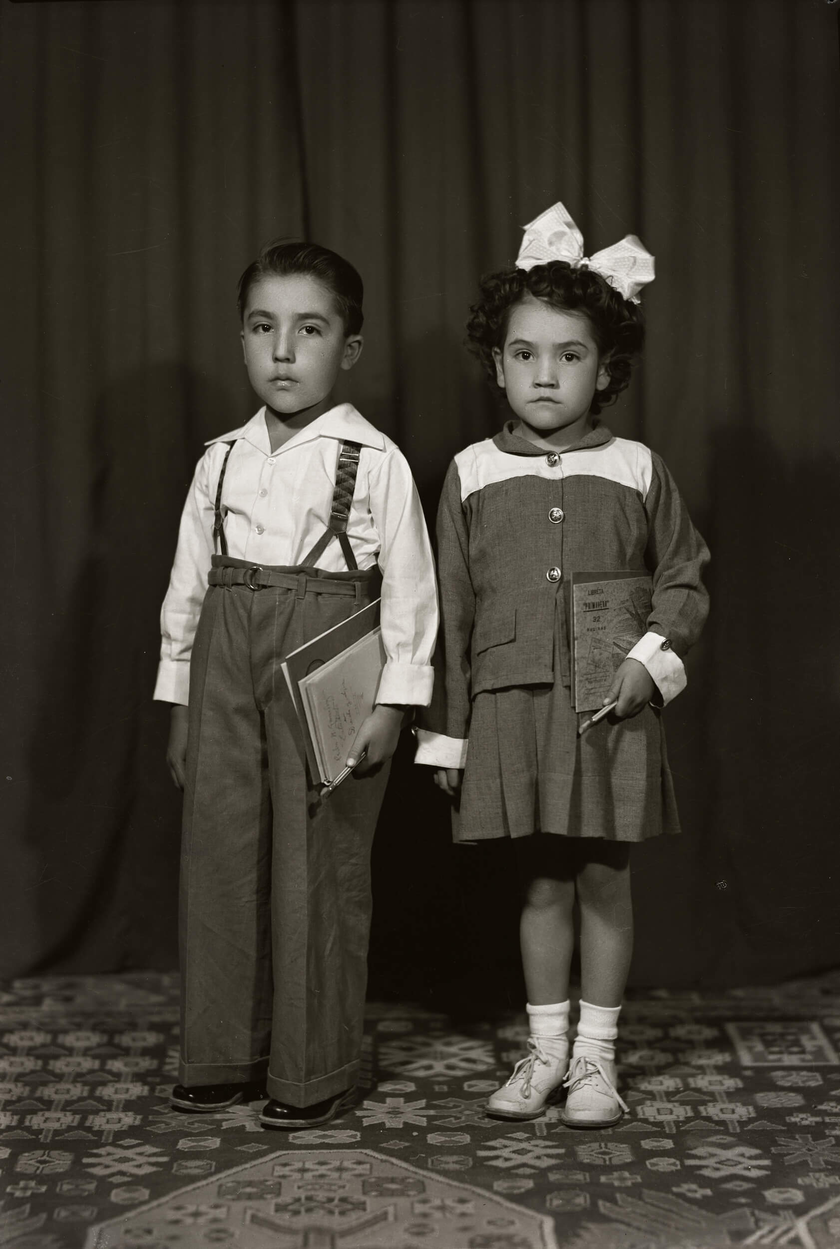 25152. Retrato de dos hermanitos de cuerpo entero con útiles escolares. La niña porta una "Libreta Primavera de 32 páginas", entre ellos, en estudio